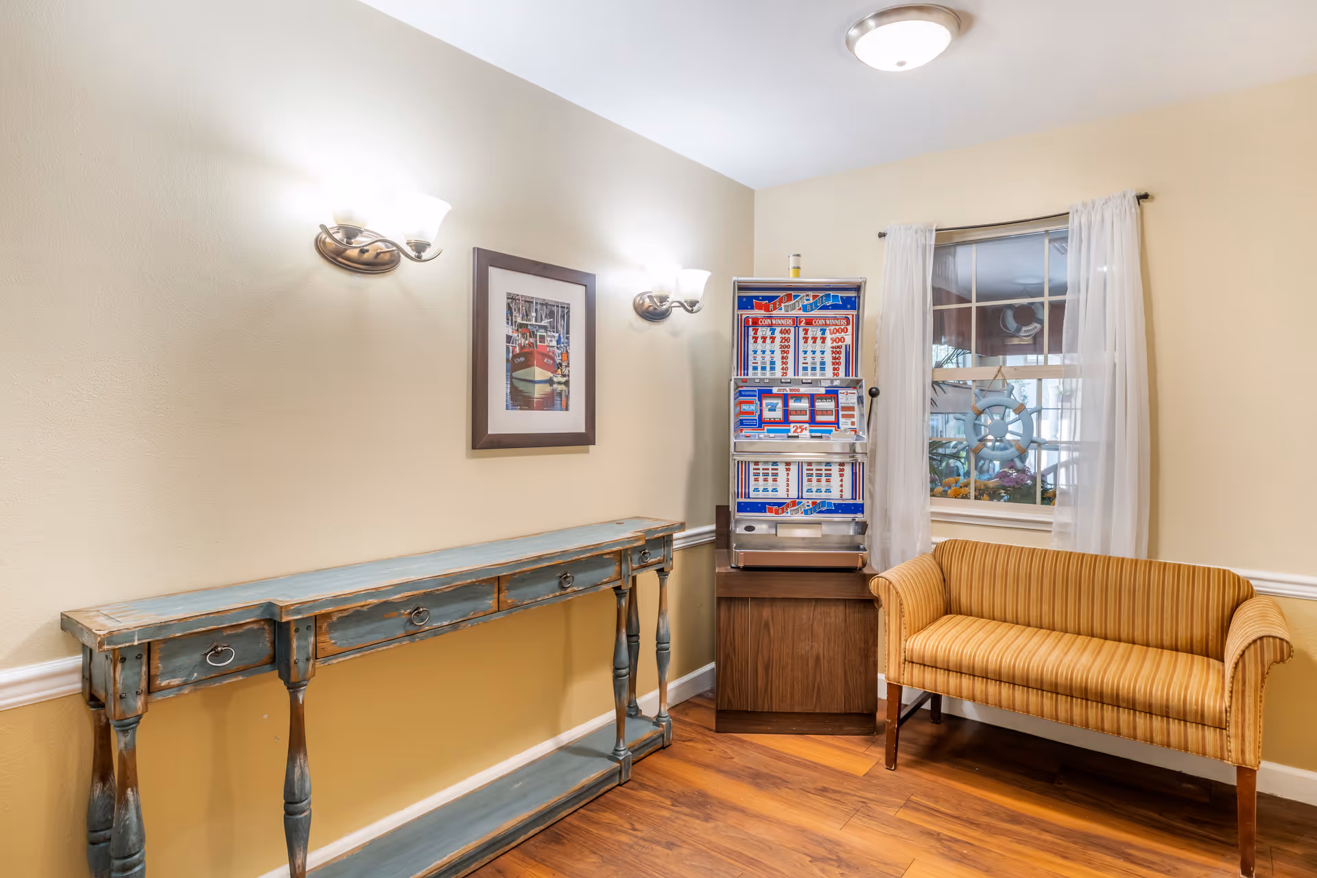 A cozy corner of a room with a vintage slot machine on a wooden stand, a striped yellow loveseat, a distressed blue console table with drawers, a framed picture of a red boat on the wall, two wall-mounted light fixtures, and a window with sheer white curtains.