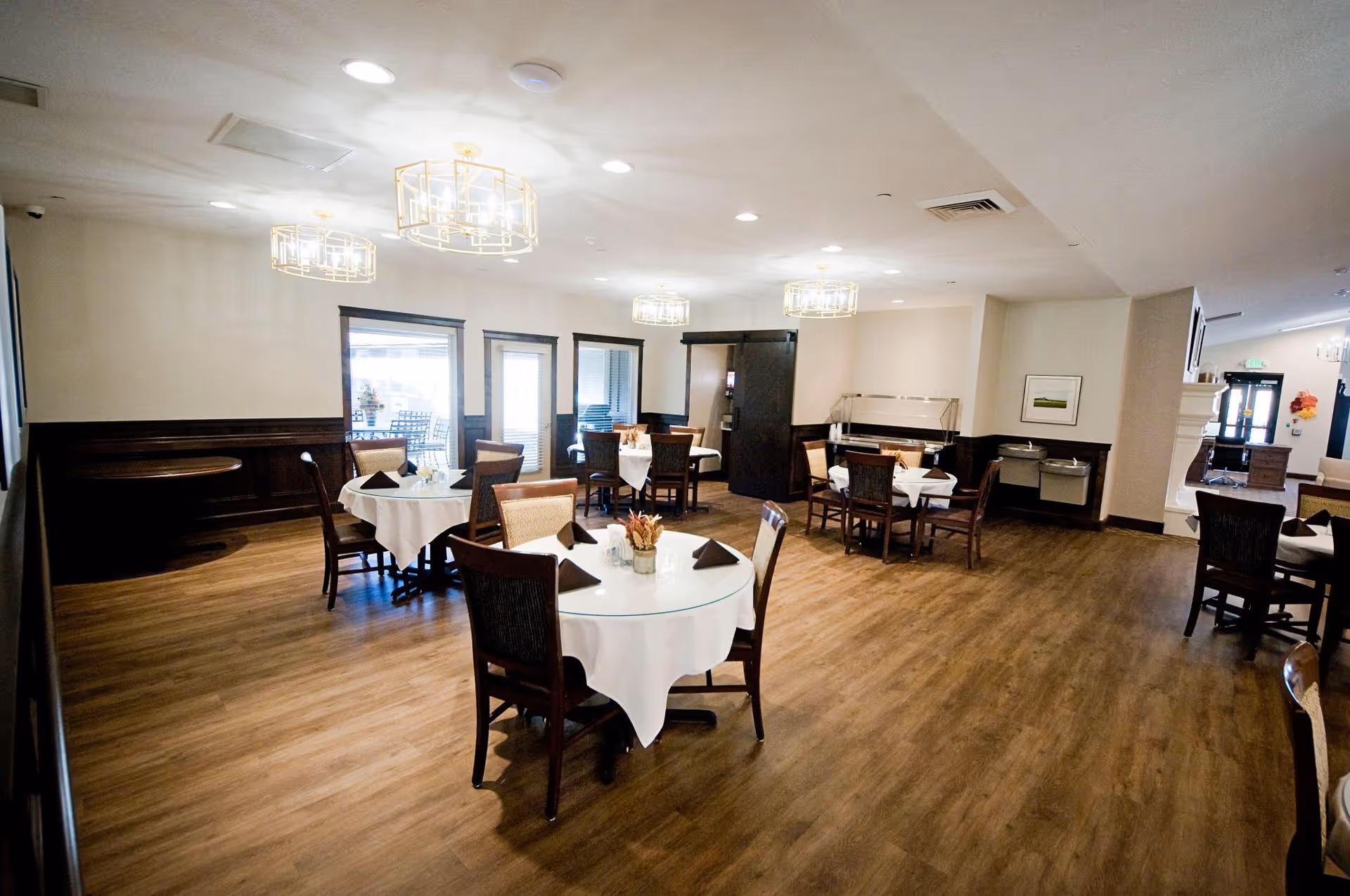 A spacious dining room with several round tables covered with white tablecloths and set with dark napkins. The room has wooden flooring, modern ceiling lights, and large windows allowing natural light. Chairs are arranged around the tables, and there is a buffet area along one wall.
