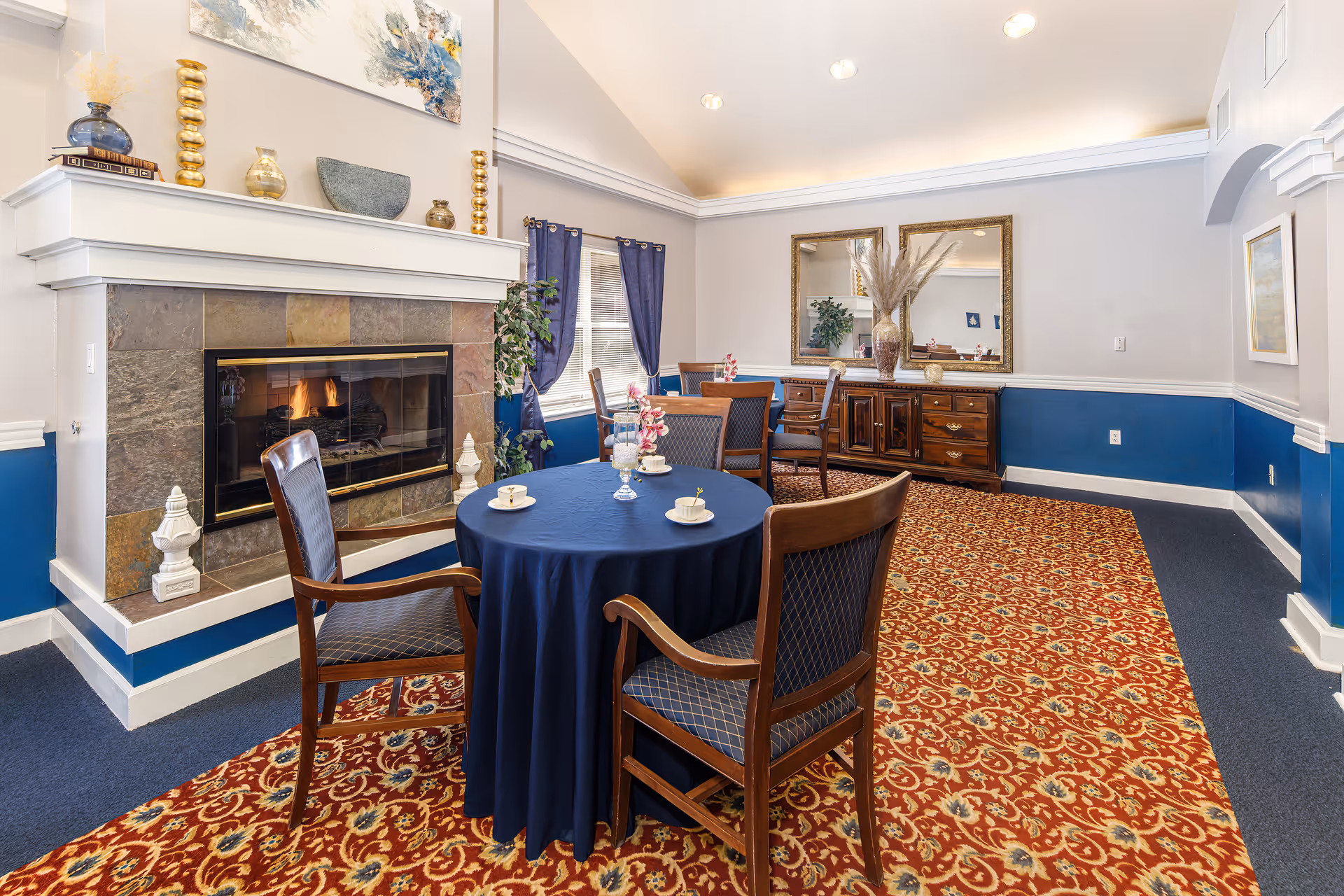 A cozy interior room with a lit fireplace surrounded by stone tiles. In front of the fireplace is a round table covered with a dark blue tablecloth, set with teacups and saucers, and surrounded by wooden chairs with blue patterned cushions. The room has a patterned red and gold carpet, blue and white walls, a large mirror above a wooden sideboard, and windows with blue curtains letting in natural light.