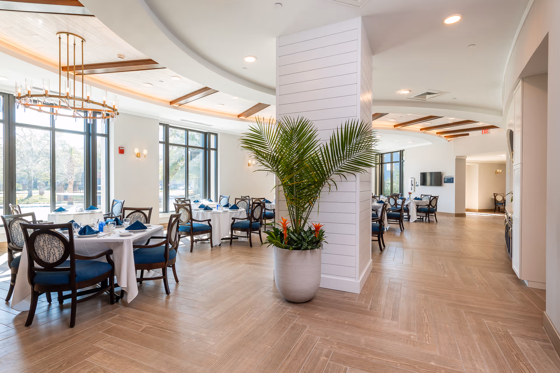 Bright and spacious dining room with large windows letting in natural light. Several round tables are set with white tablecloths, blue napkins, and dining ware. Elegant wooden chairs with blue cushions surround the tables. A large potted plant is placed near a white paneled column in the center of the room. The ceiling features wooden beams and modern light fixtures.