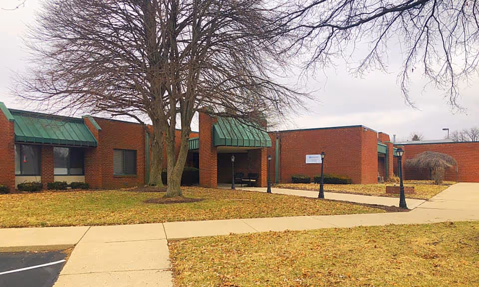 Exterior view of a single-story brick building with green metal awnings over the windows and entrance. There is a large leafless tree in the front yard, a concrete walkway leading to the entrance, and several black lamp posts along the path. The grass is brown, indicating a winter or late fall season.