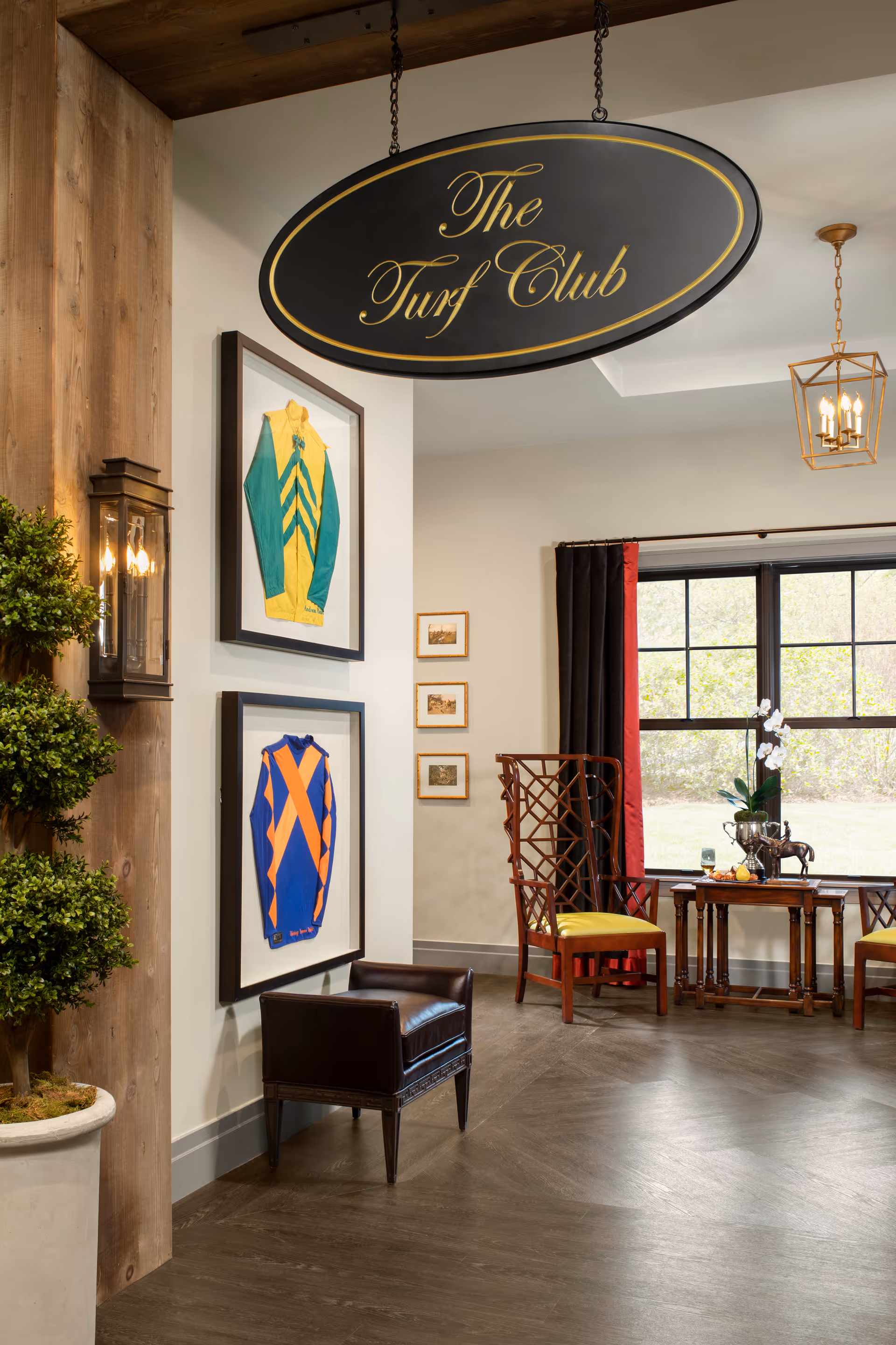 Interior view of a room with a hanging sign that reads 'The Turf Club'. The room features framed colorful sports jerseys on the wall, a small leather bench, a wooden chair with a yellow cushion, a table with decorative items including a plant and a horse figurine, and large windows with black and red curtains letting in natural light.