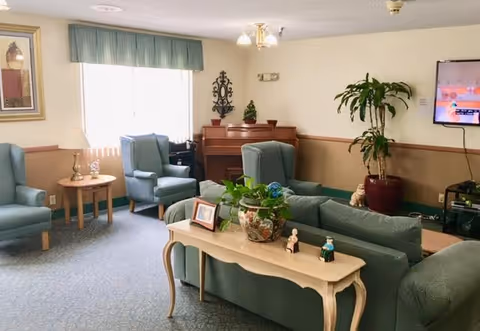 A cozy living room area in an assisted living facility with light blue armchairs and sofas arranged around a wooden table. There is a piano against the wall, a large potted plant, a wall-mounted TV, and decorative items including small figurines and framed photos on a console table. Natural light comes through a window with vertical blinds and a green valance.