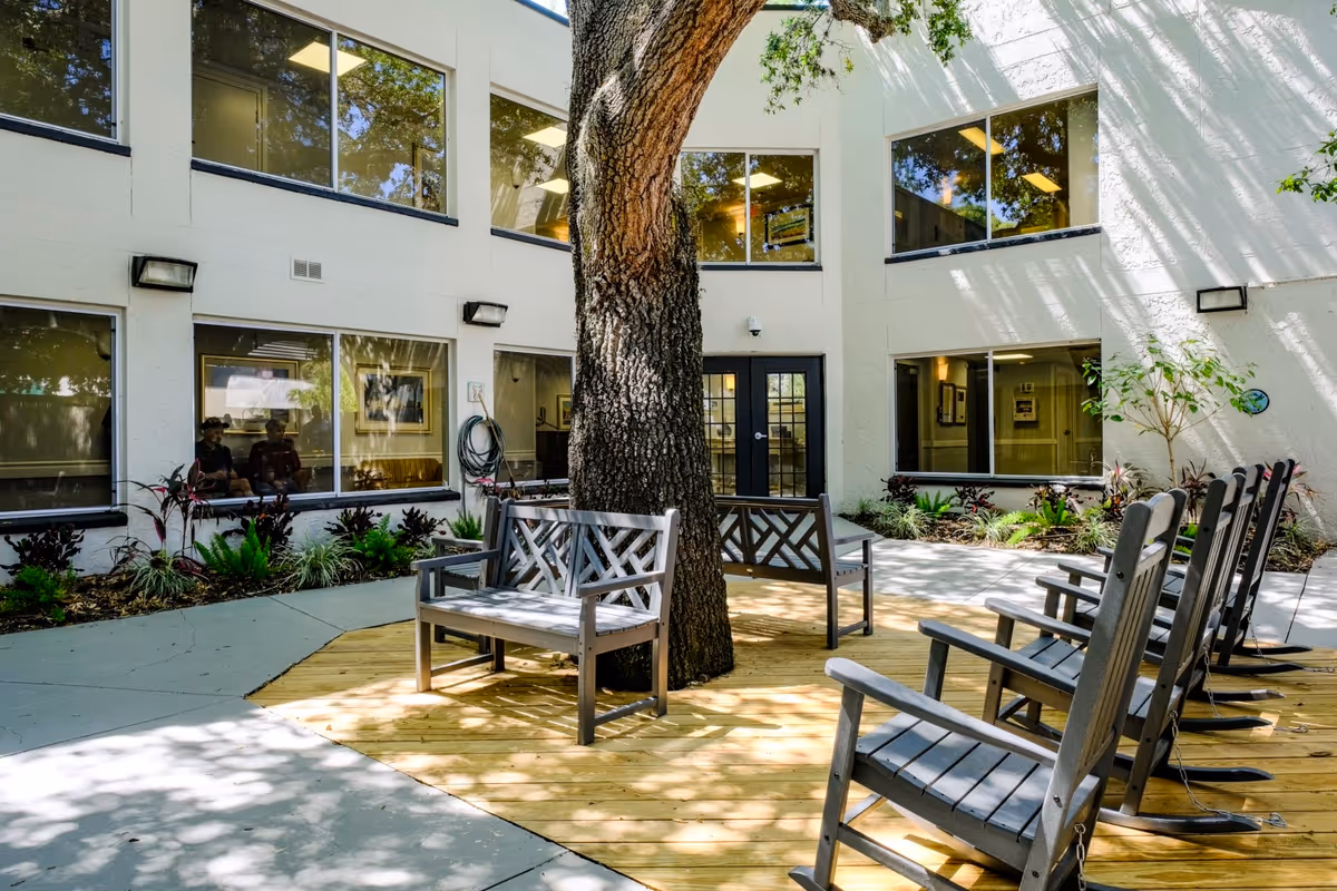 Courtyard with wooden benches and rocking chairs arranged around a large tree in front of a two-story building with windows.