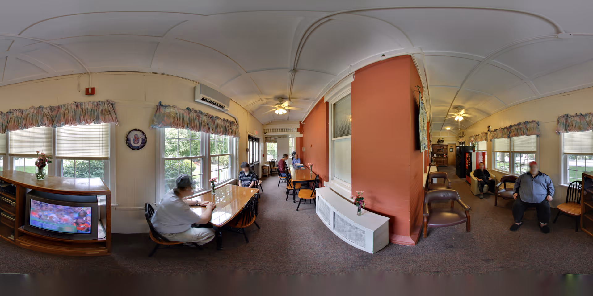 A common area in a residential care home with several elderly people seated at tables and chairs. The room has large windows with floral valances, a television on a wooden stand, ceiling fans, and a warm, inviting atmosphere. There are flowers on the tables and a red accent wall dividing the space.