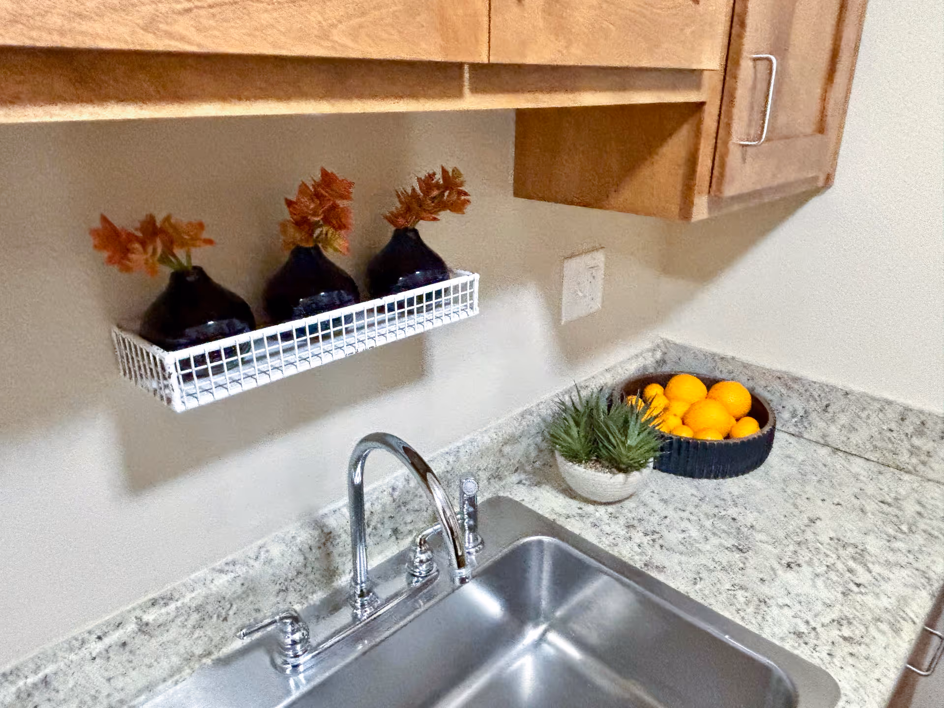 A kitchen countertop with a stainless steel sink and chrome faucet. Above the sink are wooden cabinets and a small white wire shelf holding three small dark vases with orange flowers. On the countertop next to the sink are a small potted plant and a bowl filled with oranges.