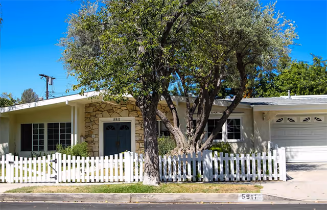 Front exterior view of a single-story building with a white picket fence, a large tree in the front yard, and a driveway leading to a garage. The building has a stone facade around the front door and several windows.