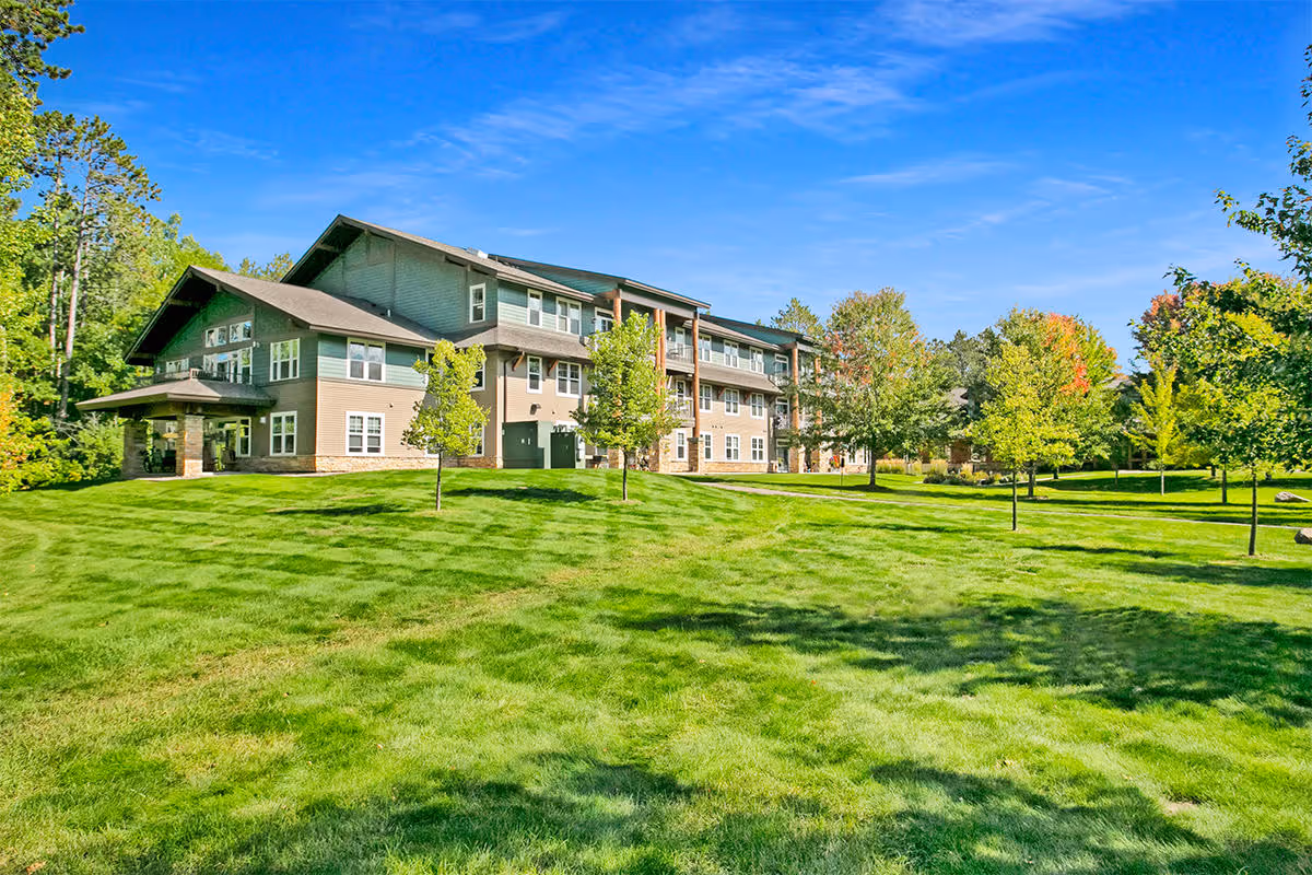 A large, multi-story residential building surrounded by a well-maintained green lawn and several trees under a clear blue sky.