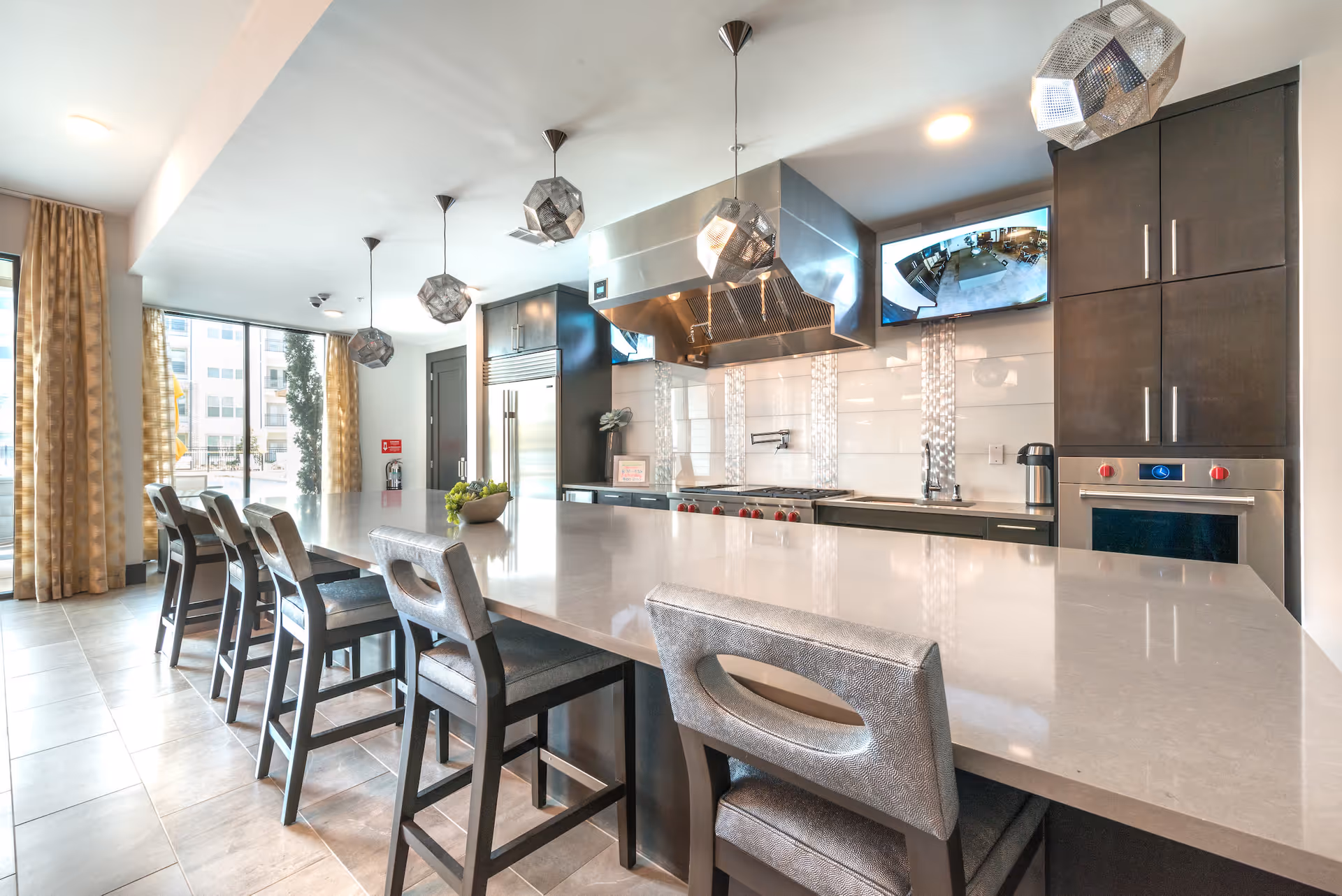 Modern kitchen area with a long island countertop and six high chairs. The kitchen features dark cabinetry, stainless steel appliances including a large refrigerator, oven, and stovetop with a vent hood. There are three geometric pendant lights hanging above the island and two wall-mounted TVs displaying security camera footage. Large windows with patterned curtains allow natural light to fill the space.
