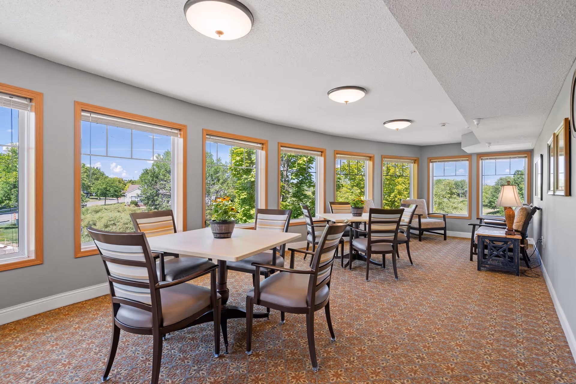 A bright and spacious common area with multiple large windows letting in natural light. The room features several tables with chairs arranged around them, each table decorated with a small potted plant. There are additional seating options including cushioned chairs and a side table with a lamp. The walls are painted light gray and the carpet has a patterned design in warm tones.