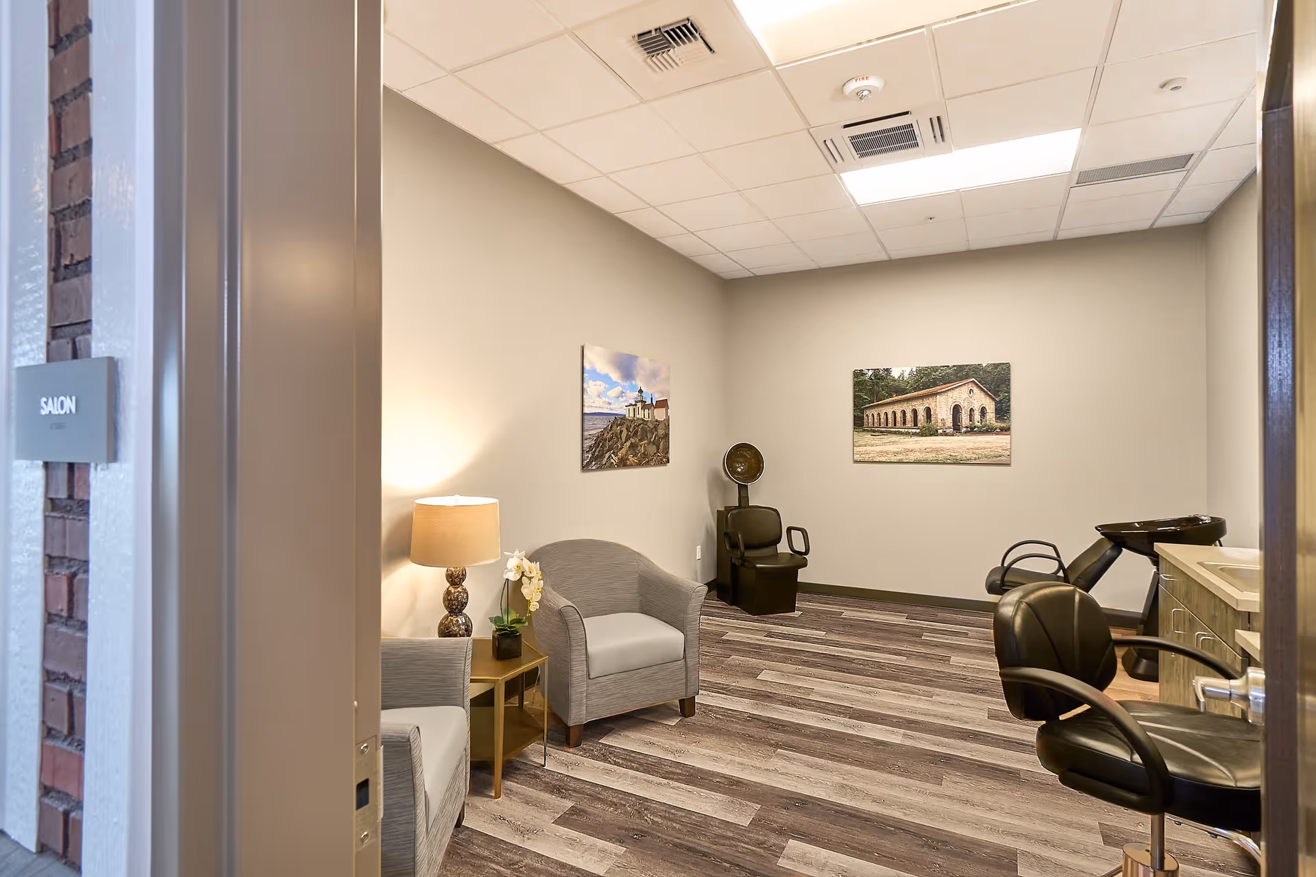 Interior salon room with armchairs, salon chairs and a hair-washing station, side table with lamp and wall artwork.