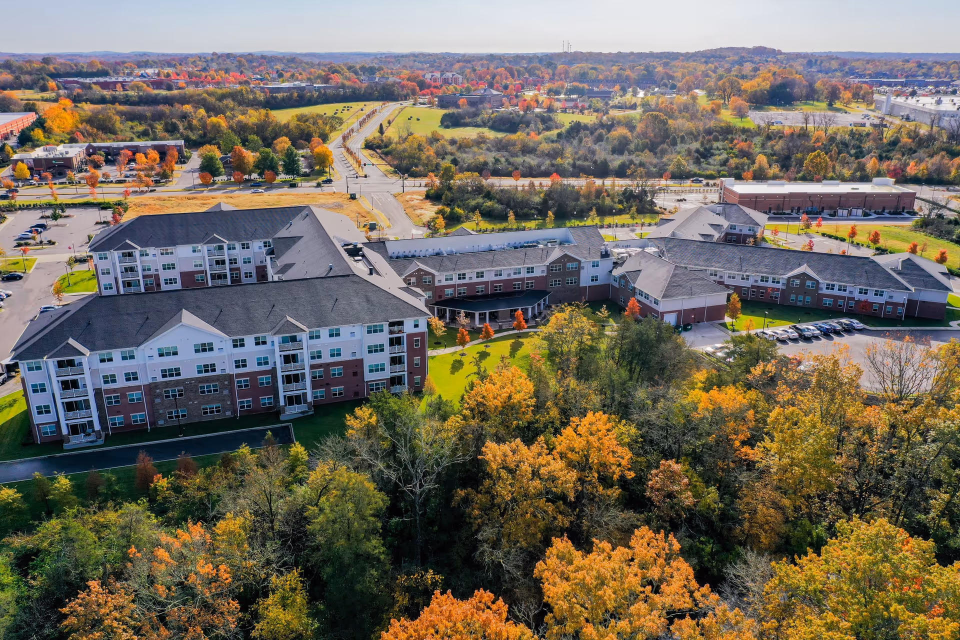 Aerial view of Clarendale at Indian Lake senior living facility surrounded by trees with autumn foliage, parking lots, and nearby roads under a clear sky.