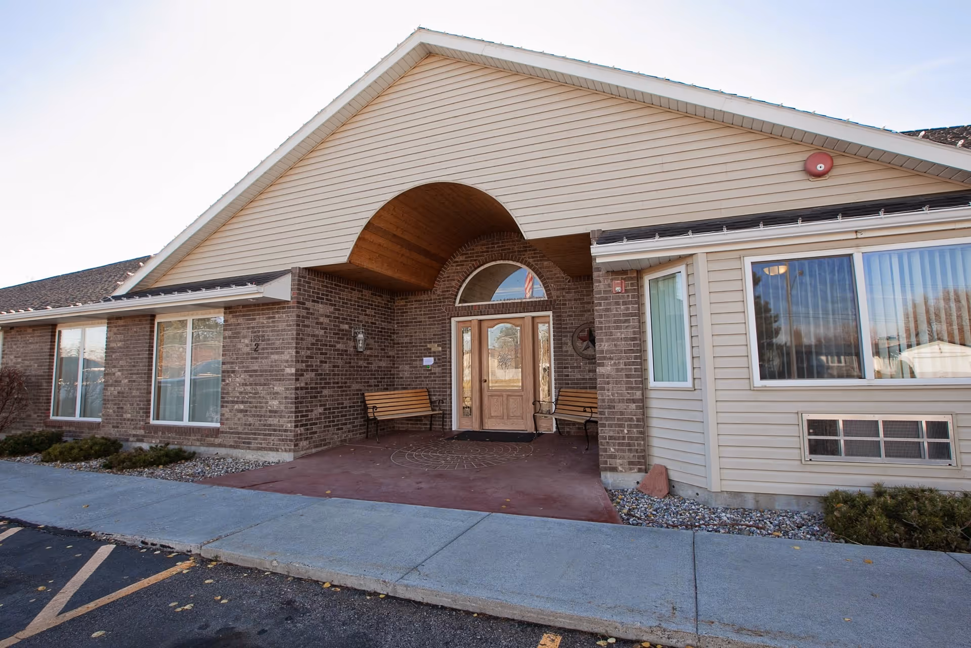 Exterior view of Turtle & Crane Assisted Living facility showing the entrance with a covered porch, two wooden benches, brick and siding walls, large windows, and a concrete sidewalk leading to the door.