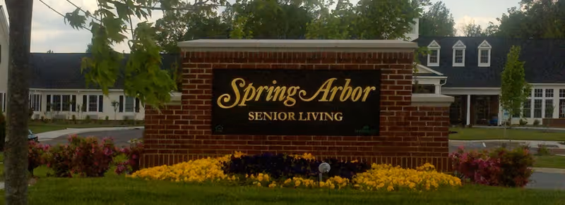 Brick sign with black background and gold lettering that reads 'Spring Arbor Senior Living' in front of a landscaped area with yellow and purple flowers, with a senior living facility building visible in the background.
