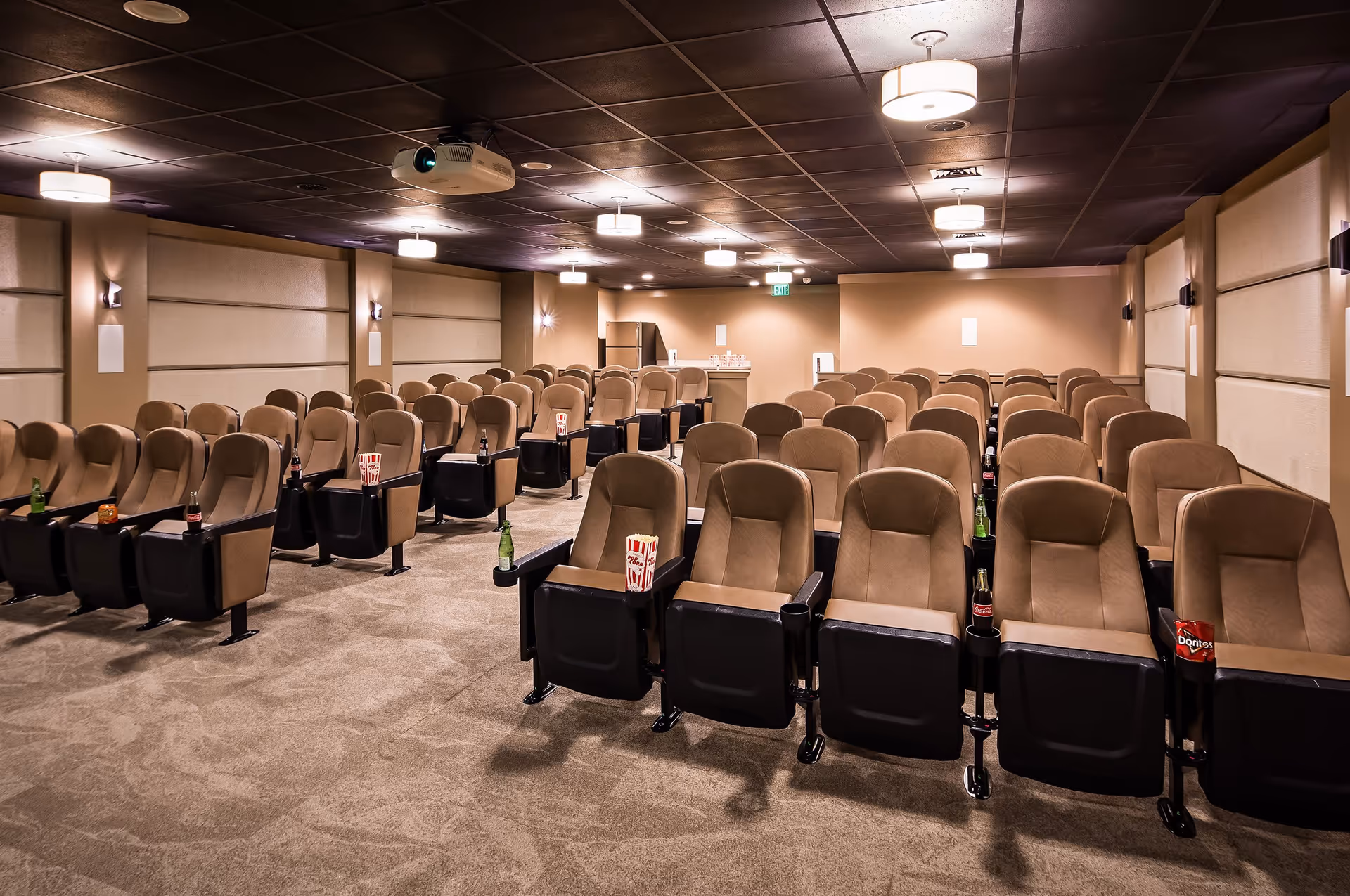 A cozy movie theater room with multiple rows of comfortable brown reclining seats, each equipped with cup holders containing popcorn, soda bottles, and snack bags. The room has a dark ceiling with several ceiling lights and a projector mounted in the center. The walls are beige with padded panels and sconces, and there is a small counter with a refrigerator and snacks at the back.