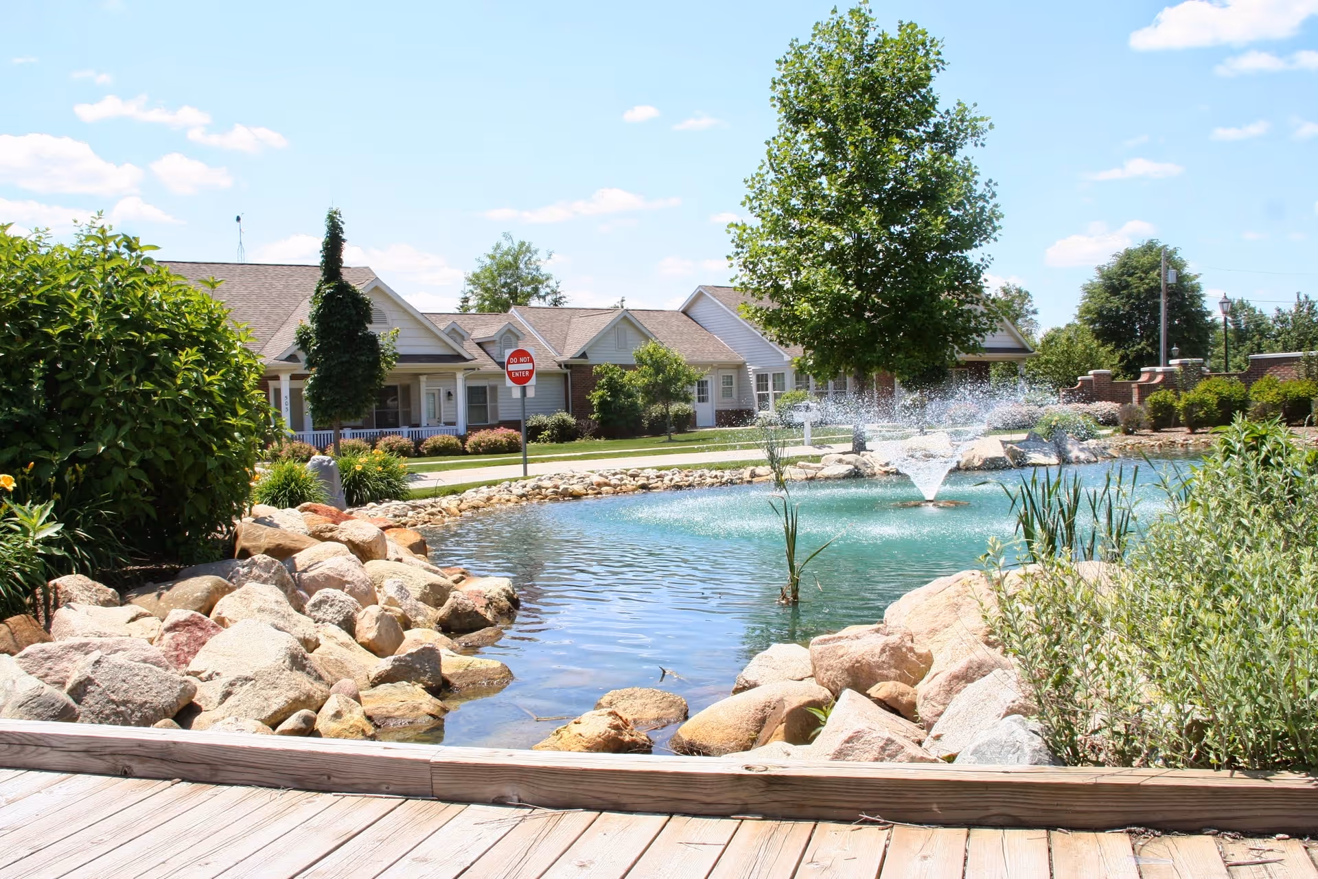 A landscaped pond with a central fountain, rock borders and greenery in front of single-story residential buildings on a sunny day.