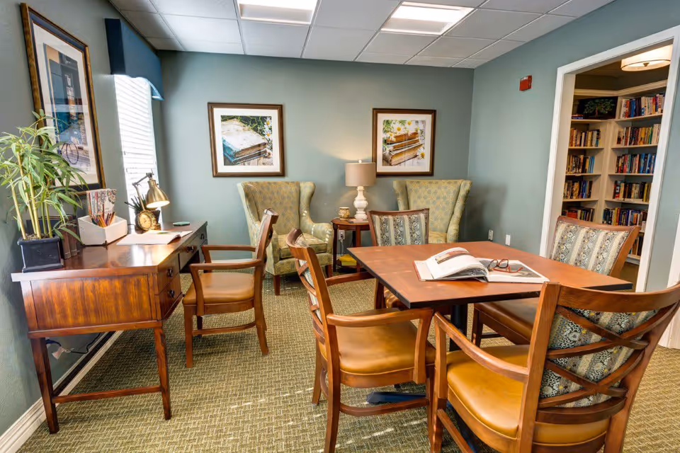 A bright common room with a wooden table and chairs, a writing desk, upholstered armchairs, framed artwork, a lamp, and a bookshelf.