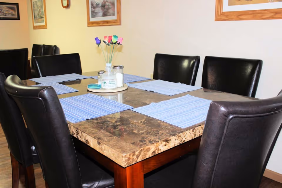 A dining table with a marble top surrounded by six black leather chairs. The table has blue striped placemats and a centerpiece with colorful artificial flowers and condiment containers. The background shows framed pictures on a light-colored wall.