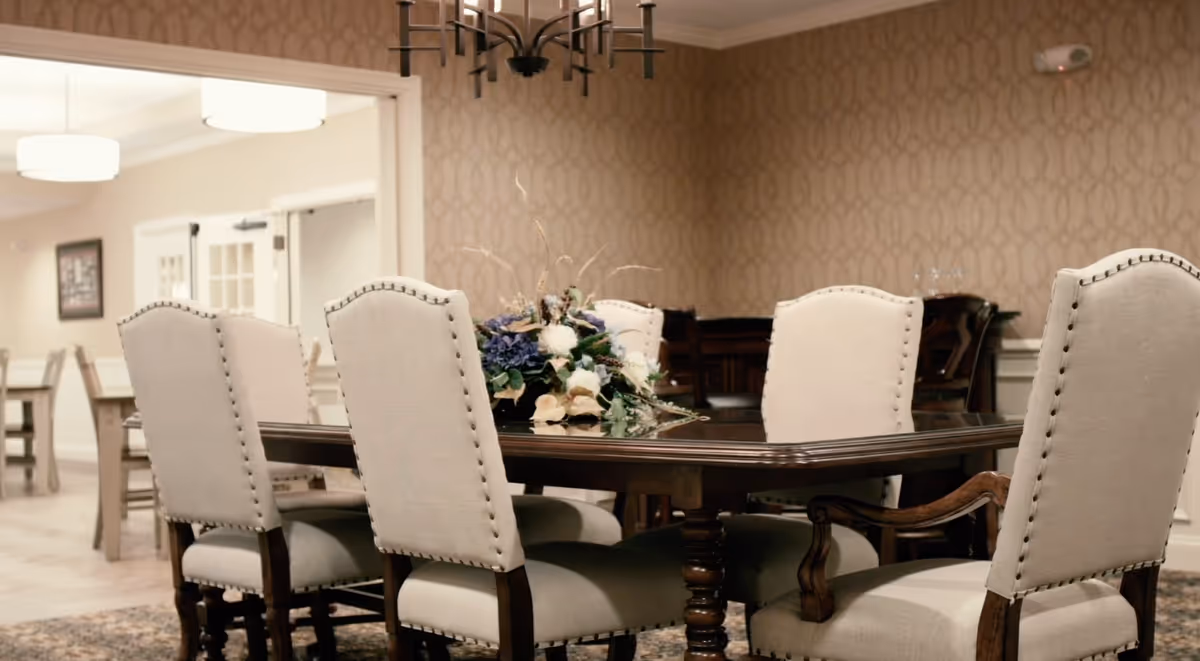 A dining room with a dark wooden table surrounded by six upholstered chairs with nailhead trim. A floral centerpiece decorates the table. The room has patterned wallpaper and a chandelier hanging above the table. In the background, there are more tables and chairs and a doorway leading to another room.