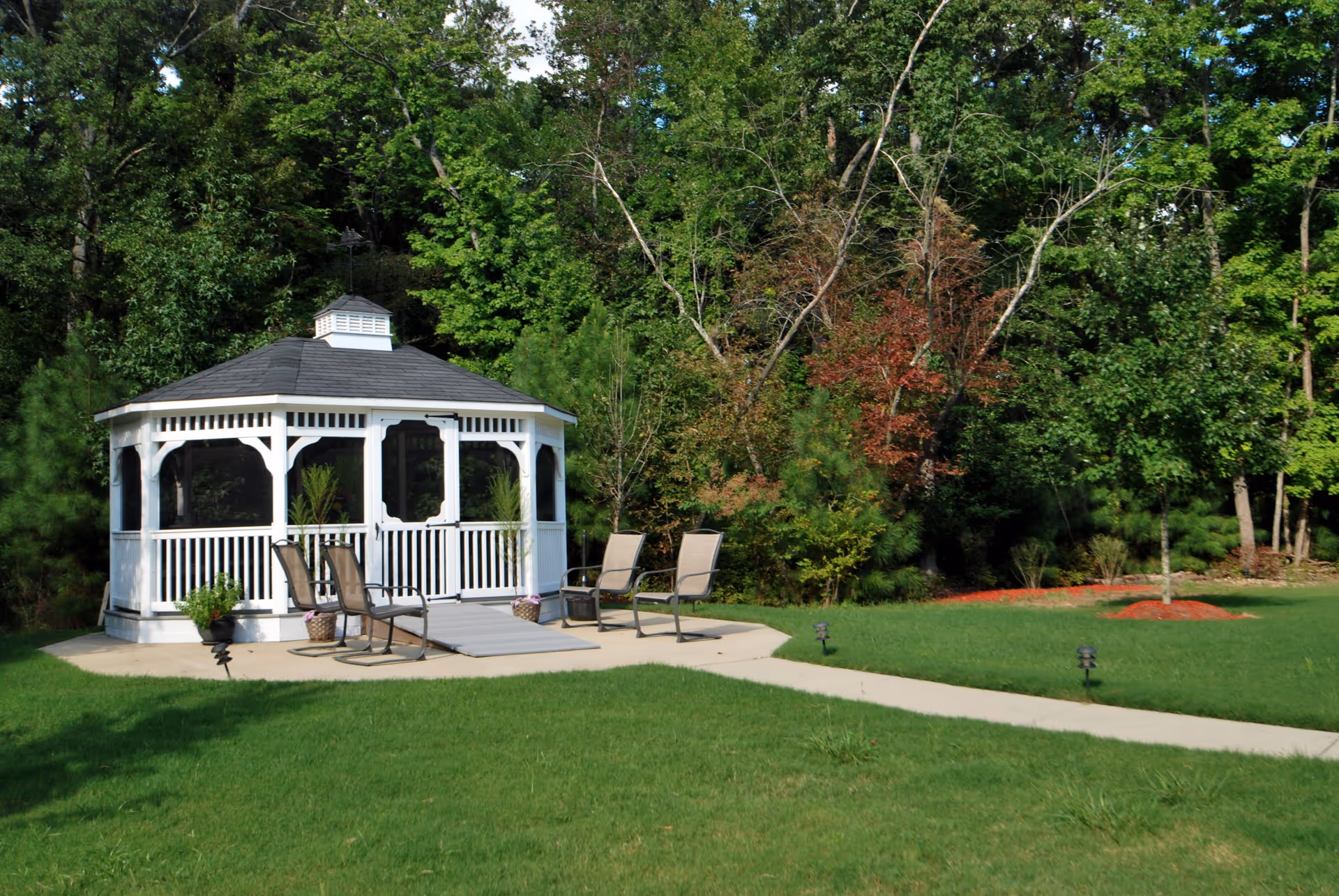 A white gazebo with a dark roof situated on a concrete patio surrounded by green grass and trees. Four outdoor chairs are placed around the gazebo, and there are potted plants near the entrance. A paved walkway leads to the gazebo through a well-maintained lawn.