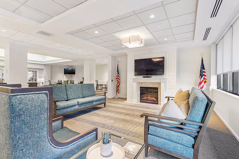 A bright and spacious lounge area in a senior living facility with blue upholstered sofas and armchairs arranged around a glass coffee table. The room features a fireplace with a mounted flat-screen TV above it, two American flags on either side of the fireplace, large windows with white blinds, and a patterned carpet. In the background, there are additional seating areas and tables.