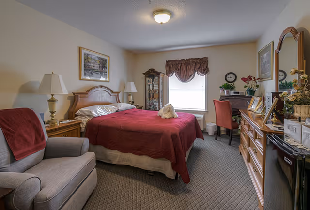 A cozy bedroom with a large bed covered in a red quilt and white pillows. A small white stuffed animal is placed on the bed. To the left of the bed is a wooden nightstand with a lamp and a gray armchair with a red towel draped over the back. A wooden dresser with a mirror and various decorative items is on the right side of the room. A window with a valance lets in natural light, and a small desk with a red chair and plants is positioned beneath the window. The room has carpeted flooring and neutral-colored walls with framed artwork.