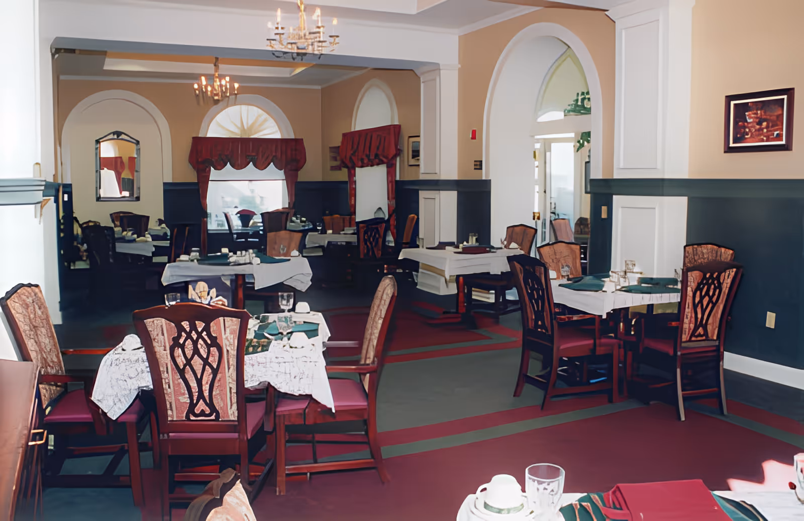 Interior view of a dining room with multiple tables and chairs arranged neatly. The tables are covered with white tablecloths and set with glasses, napkins, and utensils. The room features arched windows with red valances, chandeliers hanging from the ceiling, and walls painted in beige and dark green with white trim. A framed picture is visible on one wall.