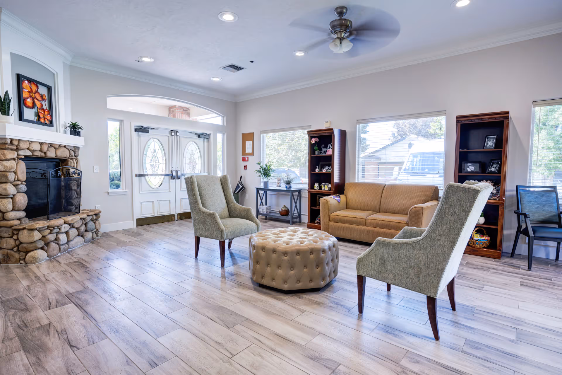 A bright and spacious senior living facility common area with a stone fireplace on the left, two green upholstered armchairs, a tan sofa, a round tufted ottoman in the center, and large windows letting in natural light. There are wooden shelves with decorative items and a ceiling fan above.