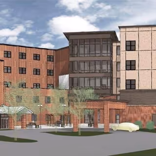 Exterior view of a multi-story nursing home building with a covered entrance, several windows, and a driveway with a parked car. The sky is partly cloudy.