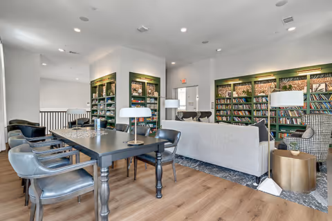 A bright and modern common area with a black table surrounded by six chairs, two white desk lamps on the table, a light gray sofa, and two armchairs. The back wall features built-in green bookshelves filled with books. The floor is a combination of wood and patterned tile, and the ceiling has recessed lighting.