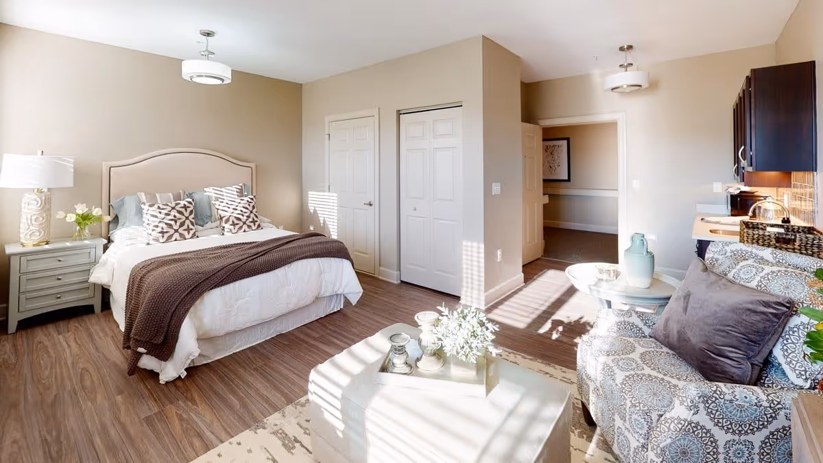 A bright and cozy bedroom with a large bed featuring patterned pillows and a brown throw blanket. There is a nightstand with a lamp and flowers on the left side of the bed. The room has wood flooring and beige walls. To the right, there is a small sitting area with a patterned armchair, a round side table with decorative items, and a kitchenette with dark cabinets and a countertop. Sunlight streams in through a window, casting shadows on the floor.