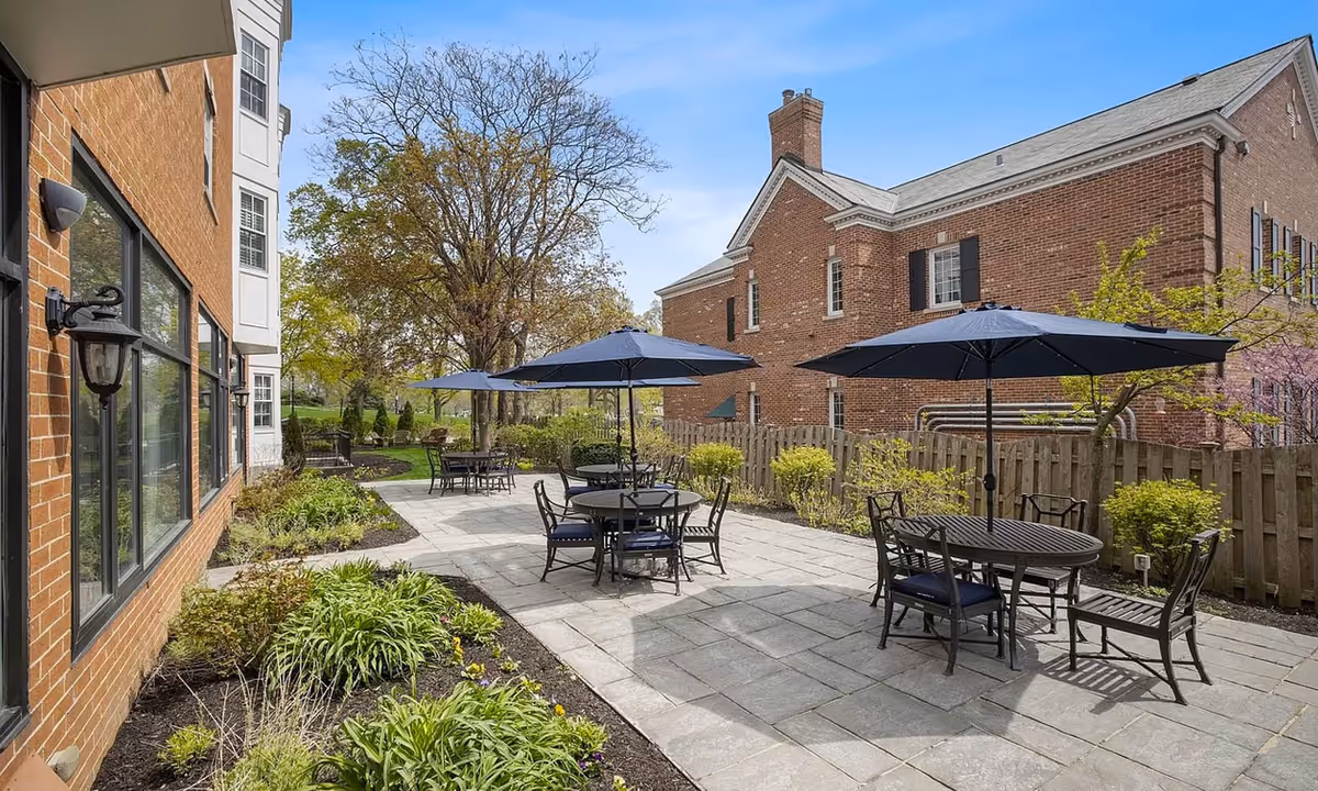 Outdoor patio with round tables, chairs, and blue umbrellas between brick buildings and landscaped garden beds.