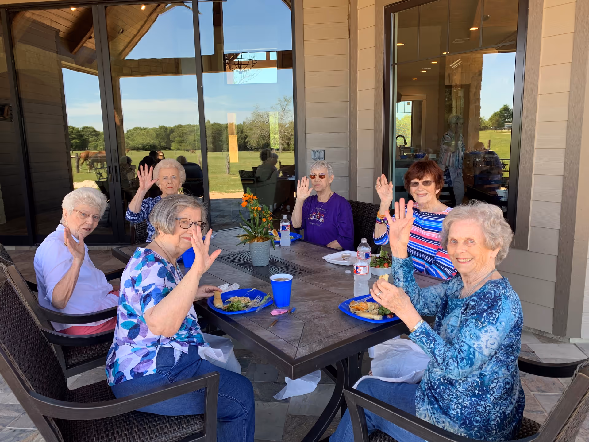 Six elderly women sitting around a rectangular outdoor table on a patio, waving at the camera. They have plates of food and drinks in front of them. Behind them is a building with large glass doors and windows reflecting the outdoor scenery of green grass and trees.