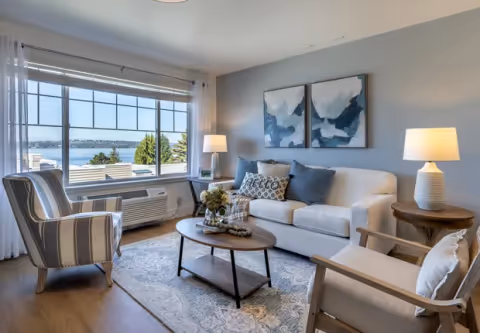 A bright and cozy living room with a large window overlooking a scenic waterfront view. The room features a white sofa with blue and patterned cushions, two armchairs, a wooden coffee table with decorative items, a patterned rug, and two table lamps on side tables. Two abstract paintings hang on the light blue wall behind the sofa.