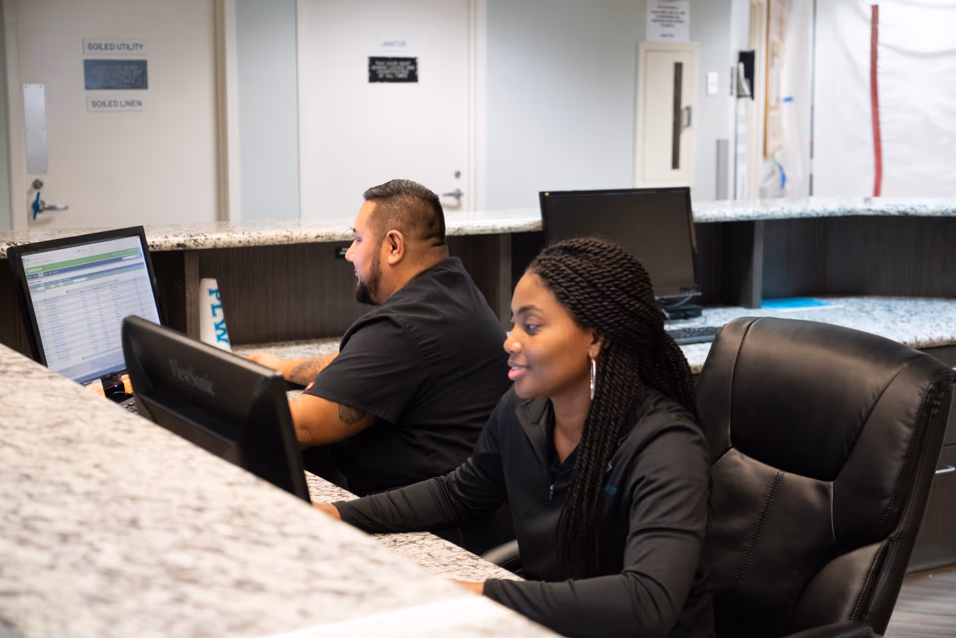 Two healthcare workers sitting at a reception desk working on computers inside a healthcare facility. The desk has a granite countertop and there are doors and signs in the background.
