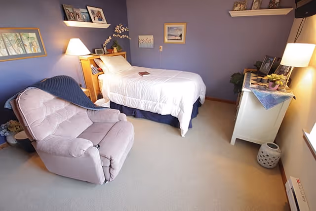 A cozy bedroom with purple walls featuring a single bed with white bedding, a wooden headboard, and a book placed on the bed. There is a comfortable pink recliner chair with a blue blanket draped over it. The room also includes a white dresser with a lamp, plants, and framed pictures on top. Shelves with decorative items and framed artwork hang on the walls.
