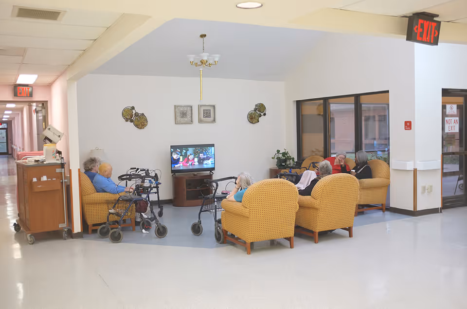 A group of elderly people sitting in a common area of an assisted living facility, watching television. The room has yellow armchairs arranged in a semi-circle, walkers nearby, and wall decorations. There is a hallway to the left and large windows on the right.