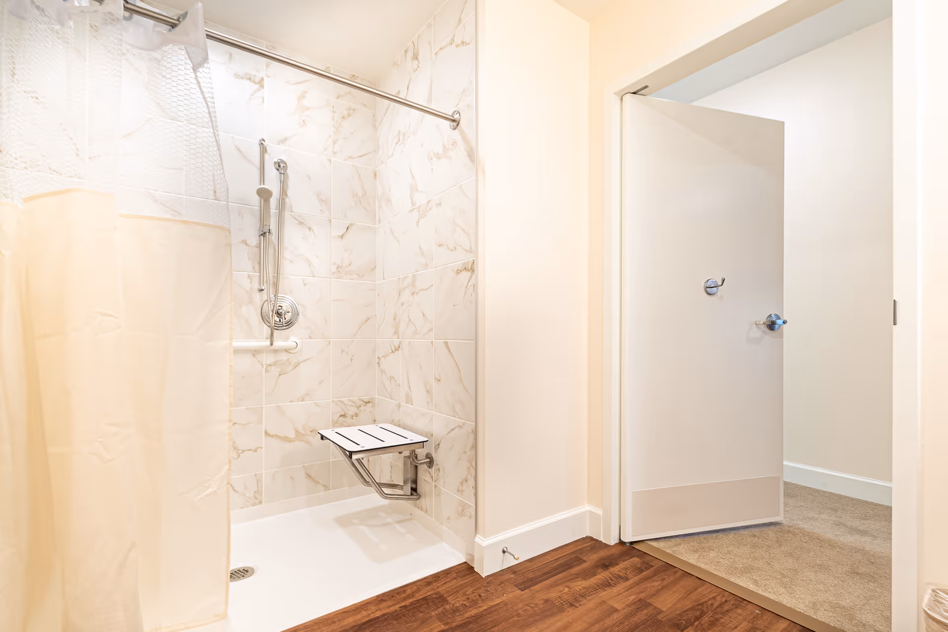 A bathroom with a walk-in shower featuring marble-patterned tiles, a foldable white shower seat, a handheld showerhead, and a beige shower curtain. The bathroom has wooden flooring and an open door leading to a carpeted room.