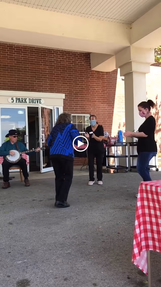 An outdoor covered area at Peachtree Village Assisted Living with a man playing a banjo seated near an open door labeled '5 Park Drive'. Two women wearing face masks are standing and interacting, while another woman in a blue patterned jacket is dancing. A table with a red and white checkered tablecloth is partially visible in the foreground.
