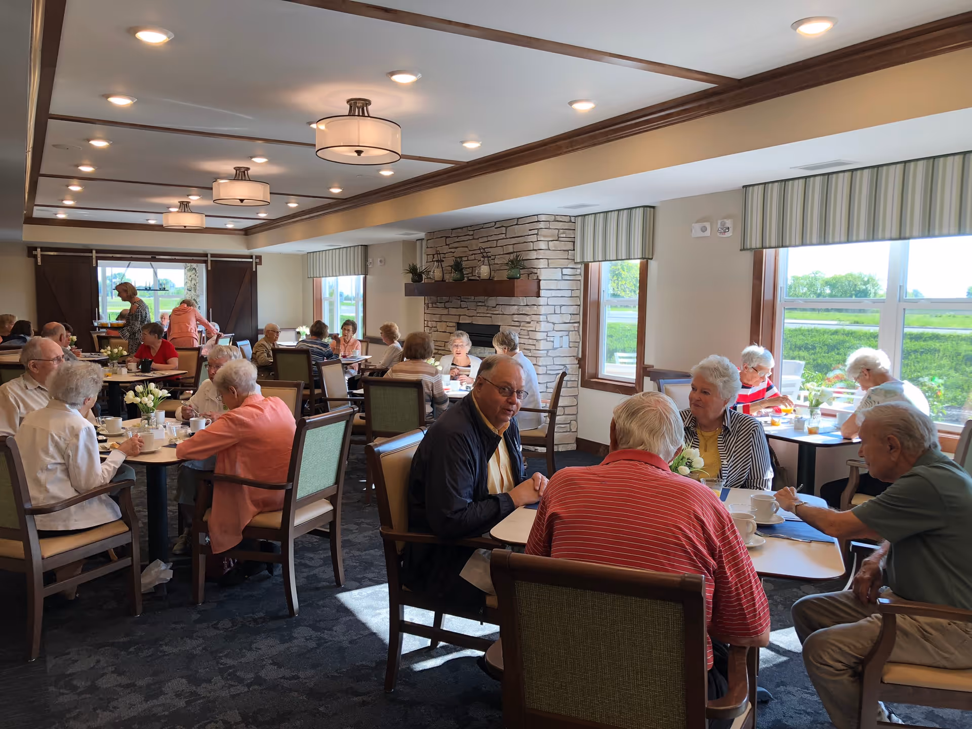 A dining room in a senior living facility with multiple elderly people sitting at tables, eating and conversing. The room has large windows with striped valances, a stone fireplace, and ceiling lights with wooden beams. The atmosphere is bright and social.
