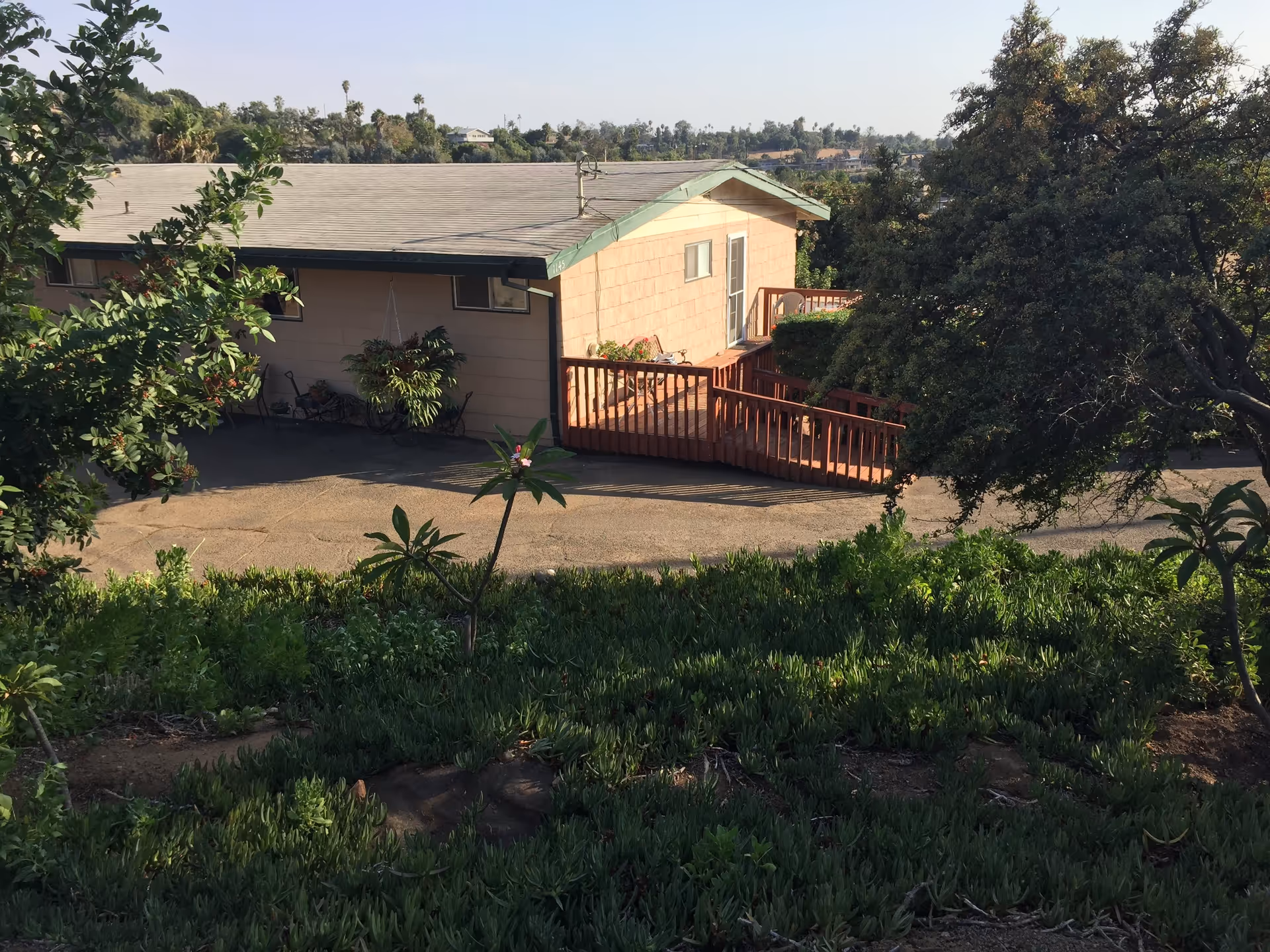 Exterior view of a single-story building with a sloped roof and a wooden ramp leading to a door. The building is surrounded by greenery including bushes, trees, and plants, with a paved area in front. The background shows a distant view of more trees and houses under a clear sky.
