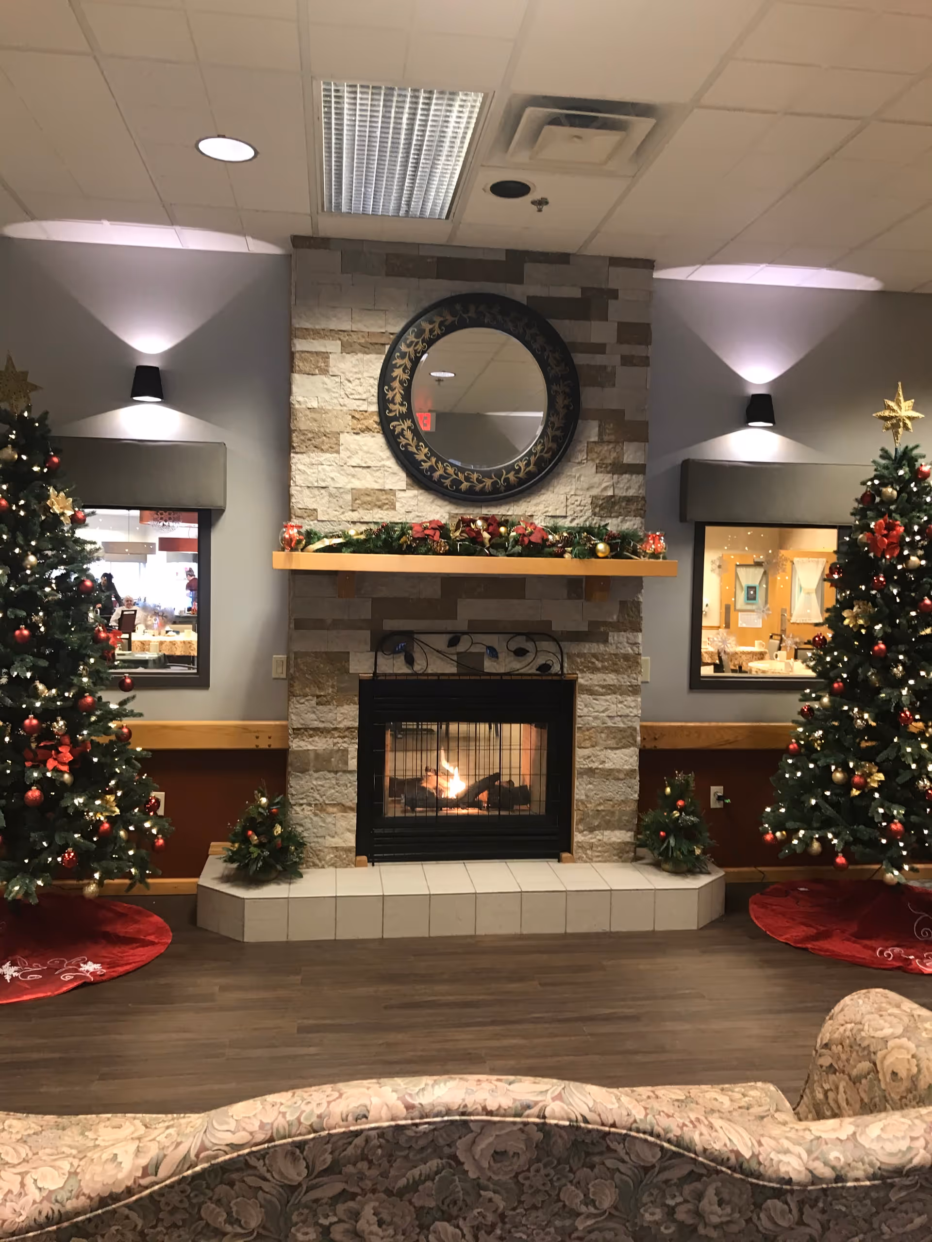 Cozy senior living room decorated for Christmas with two decorated Christmas trees on either side of a stone fireplace. The fireplace has a fire burning and a round decorative mirror above it. There are small Christmas wreaths on the hearth and a floral patterned couch in the foreground.