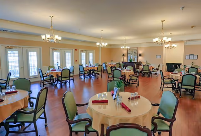 A spacious dining room with multiple round tables covered with beige tablecloths, each set with glasses and red napkins. The room has green cushioned chairs, wooden flooring, chandeliers hanging from the ceiling, and several windows and glass doors allowing natural light to enter.