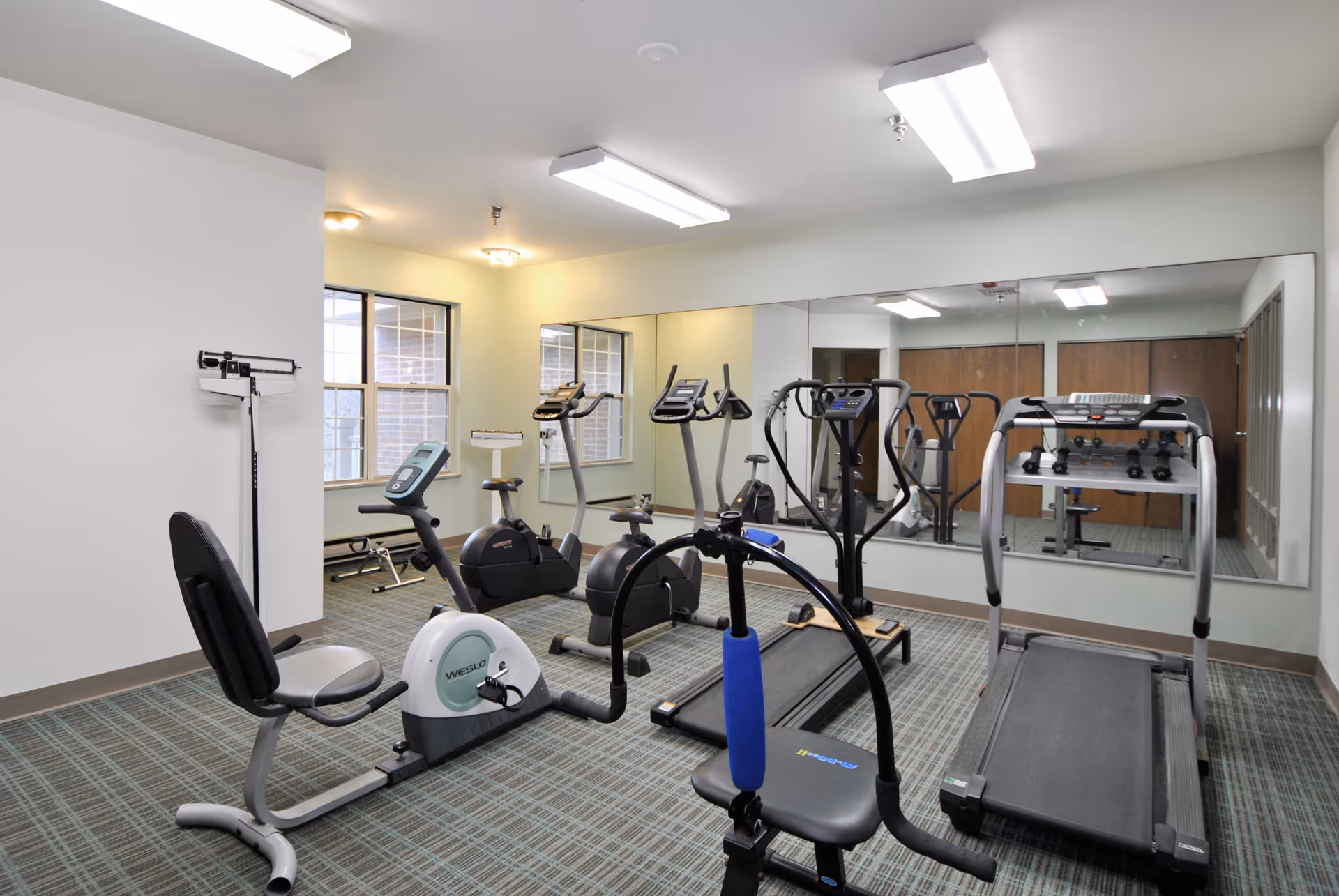 A small fitness room with exercise equipment including a recumbent bike, treadmill, elliptical machines, and a weight bench. The room has large mirrors on one wall, windows letting in natural light, and a scale in the corner.
