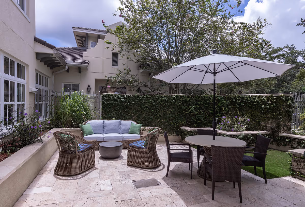 Outdoor patio area with a round table and four wicker chairs under a large white umbrella. There is also a cushioned wicker sofa with two green pillows and two matching wicker chairs around a small round coffee table. The patio is surrounded by a low wall with plants and a tall hedge, with a building visible in the background under a partly cloudy sky.