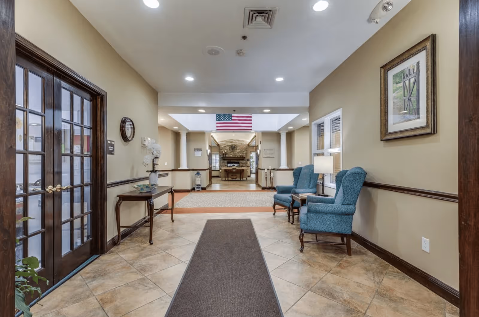 Interior view of a senior living facility hallway with beige walls and tiled floor. On the right side, there are two blue upholstered armchairs with a small table and lamp between them. A framed picture hangs above the chairs. On the left side, there is a wooden table with a decorative vase and a clock on the wall above it. At the end of the hallway, an American flag is hanging from the ceiling, and beyond it is a room with a stone fireplace and furniture. Double glass doors are visible on the left side near the entrance.
