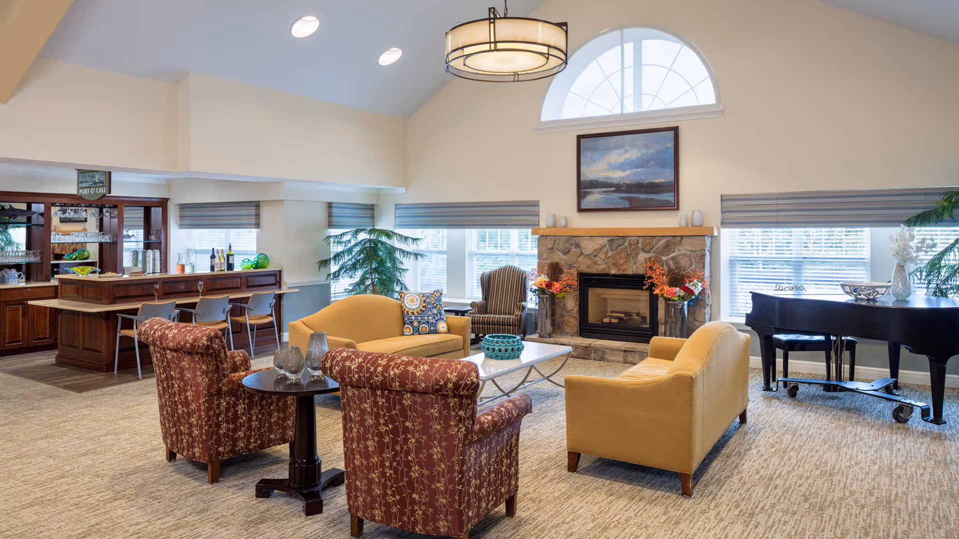 A spacious and well-lit living room area in a senior living facility featuring two yellow sofas, two patterned armchairs, a stone fireplace with a painting above it, a black grand piano, and a wooden bar area with stools. Large windows with blinds allow natural light to fill the room, and decorative plants and vases add a cozy atmosphere.