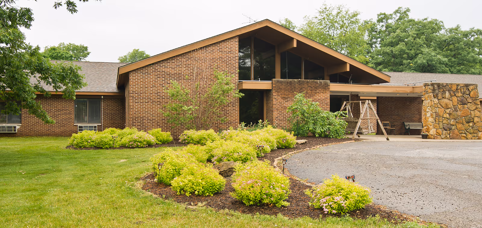 Exterior view of a single-story brick building with a sloped roof, surrounded by green grass, bushes, and trees. There is a paved driveway in front and a wooden swing near the entrance.
