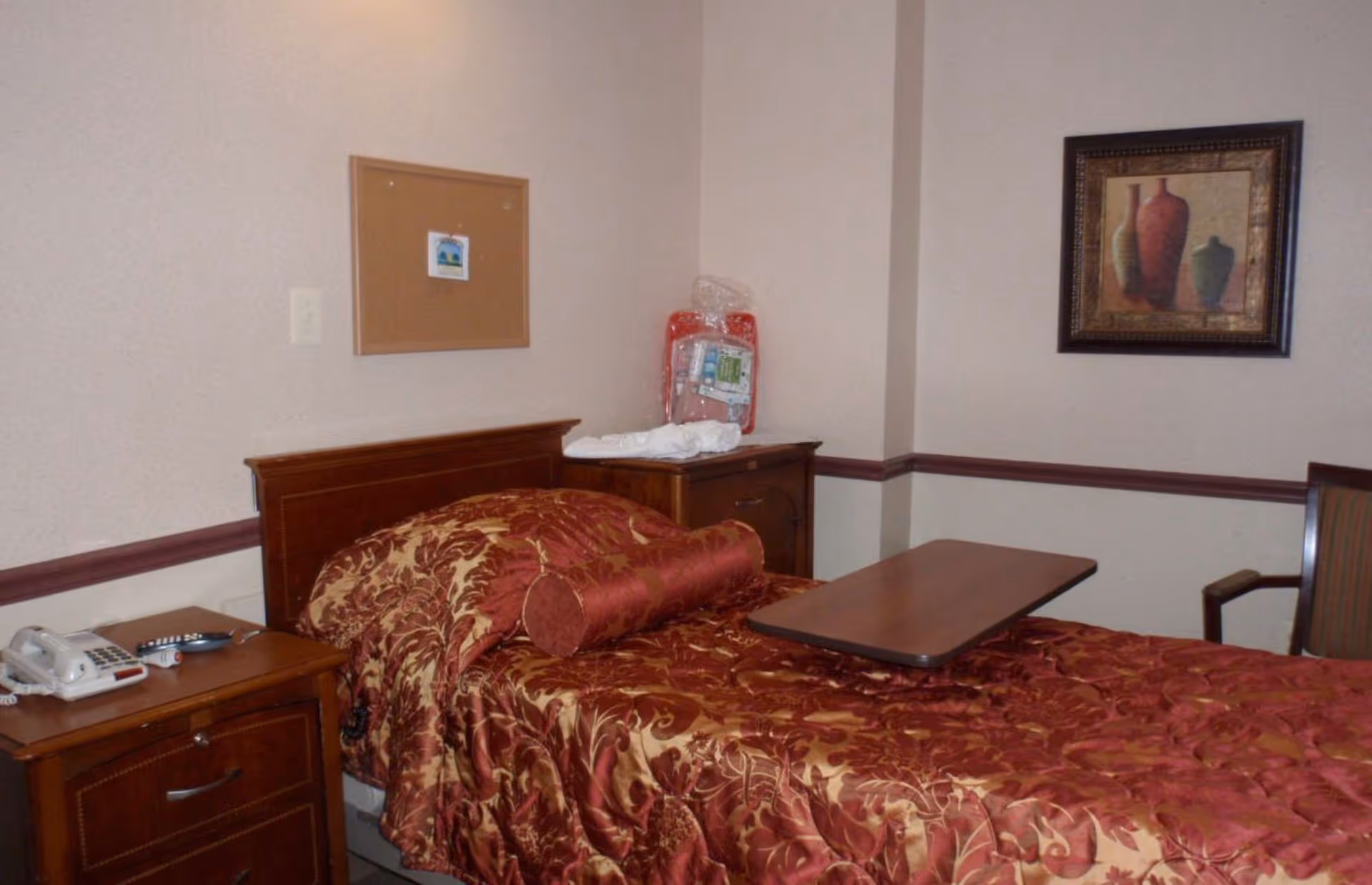A nursing facility bedroom with a single bed covered in a red and gold patterned bedspread and matching bolster pillow. There is a wooden nightstand with a telephone and remote control, a wooden cabinet with a folded towel and a packaged item on top, a chair with wooden arms, a framed painting of vases on the wall, and a cork bulletin board with a small paper pinned to it.