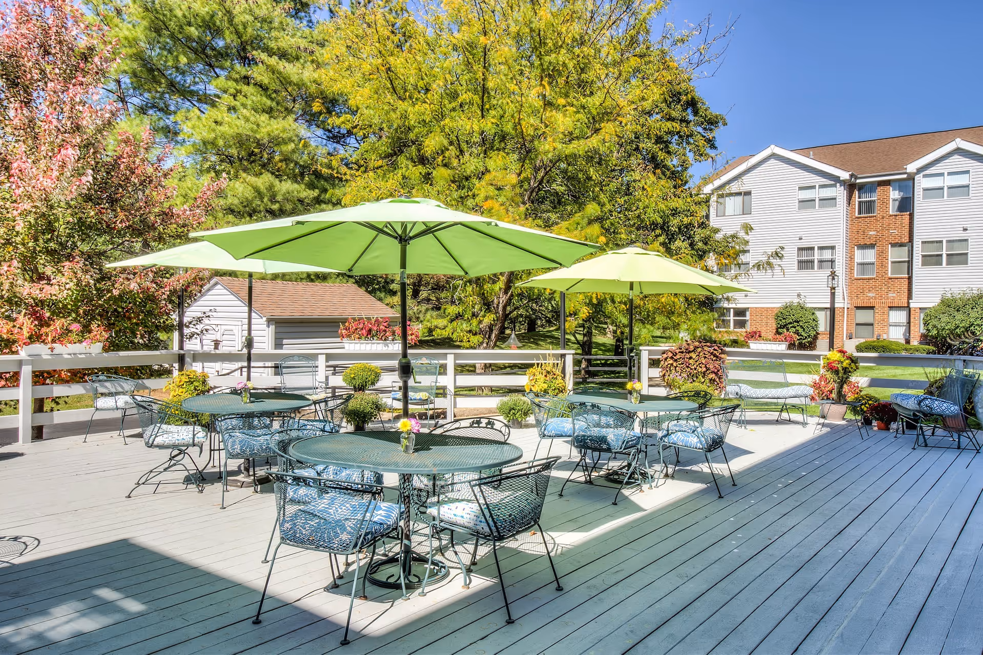 Outdoor patio area with several round metal tables and chairs, each table shaded by large green umbrellas. The patio is surrounded by a white wooden fence, with trees and shrubs in the background and a multi-story residential building visible behind the greenery.